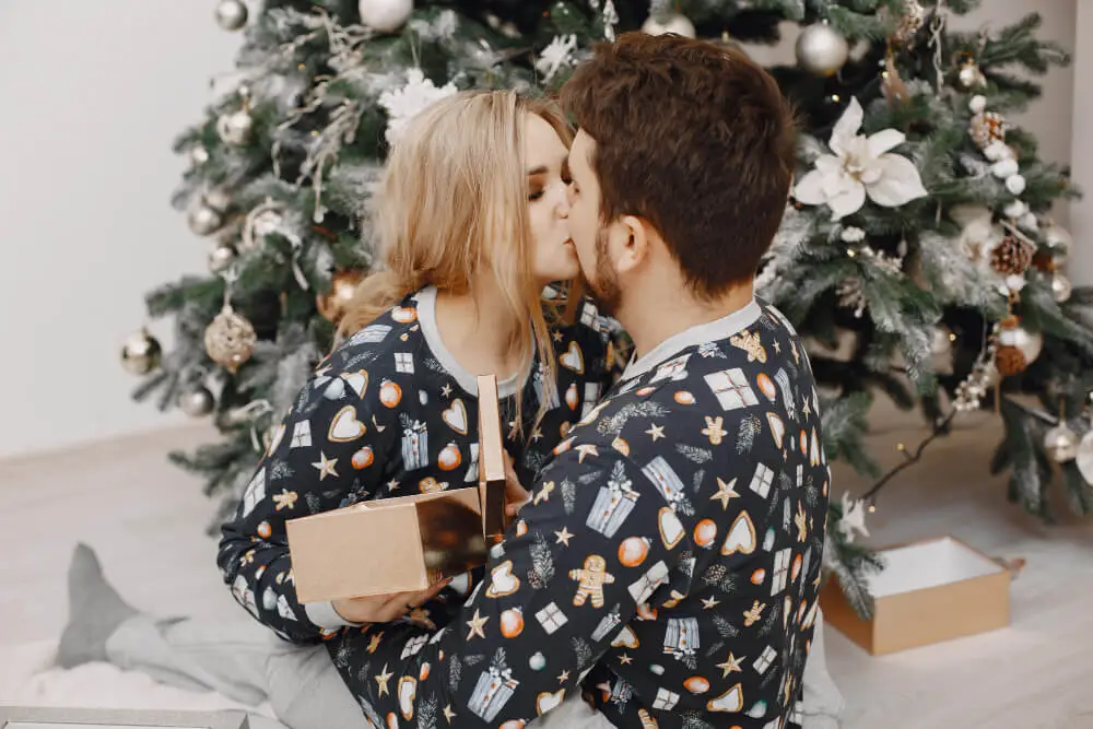A couple sitting on the floor in matching Christmas pajamas, sharing a kiss in front of a beautifully decorated Christmas tree, with a gift box in their hands