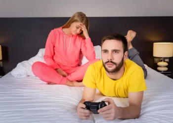Frustrated wife sitting on the bed while her husband is focused on playing video games, illustrating signs of feeling neglected in a relationship