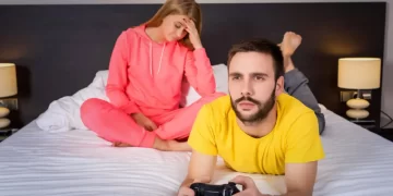 Frustrated wife sitting on the bed while her husband is focused on playing video games, illustrating signs of feeling neglected in a relationship