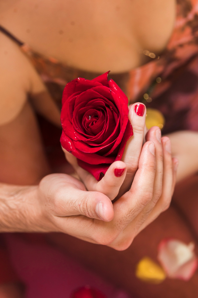 Close-up of a couple holding a single red rose together, symbolizing love and romance, with rose petals scattered around